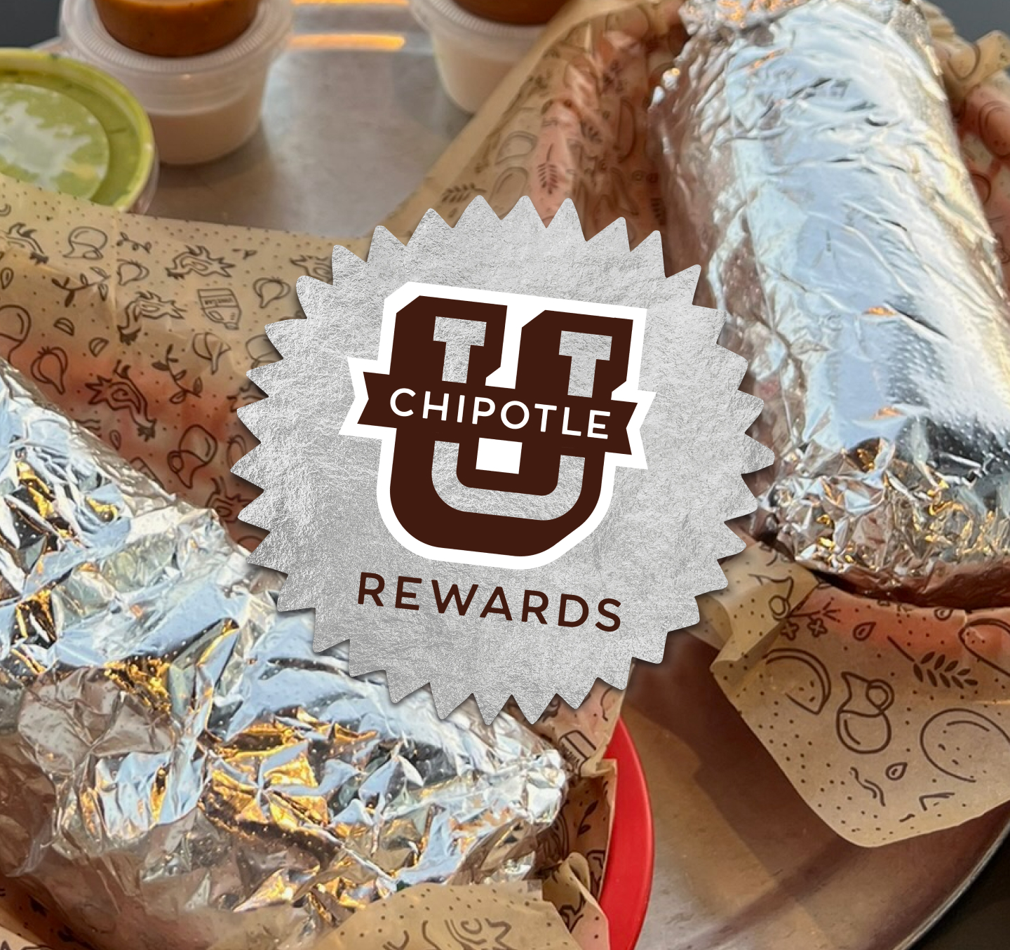 Two Chipotle Burritos wrapped in foil on a table in a Chipotle restaurant with a Chipotle U Rewards logo on top of it.